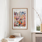 a framed poster of a colorful floral scene hanging on a wall, with a white table and a vase of flowers in the foreground.