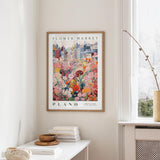 a framed poster of a colorful floral scene hanging on a wall, with a white table and a vase of flowers in the foreground.
