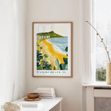 a framed poster of Waiiki Beach hanging on a wall, with a window and a small table with books and a vase in the foreground.
