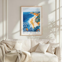 a framed poster of Bondi Beach hanging on a wall above a white couch with a blanket draped over it.