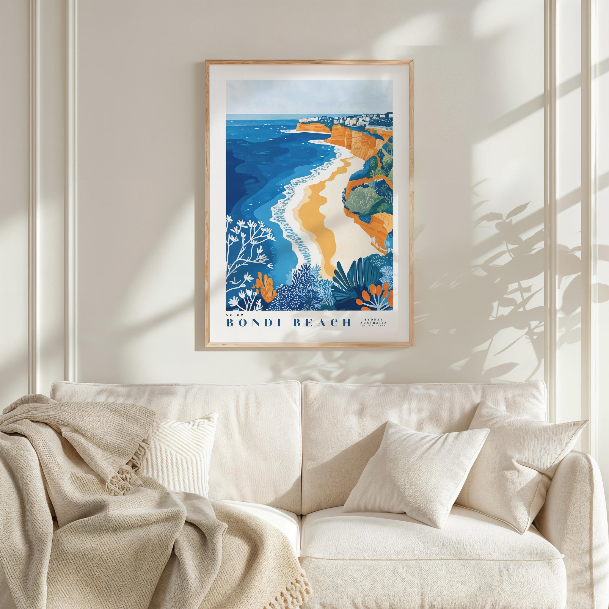a framed poster of Bondi Beach hanging on a wall above a white couch with a blanket draped over it.