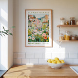 a framed poster of a flower market in Monterey, California, hanging on a wall above a kitchen counter with a bowl of lemons and jars of food on the counter.