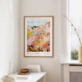 a framed poster of a colorful floral scene hanging on a wall, with a white table and a vase of flowers in the foreground.
