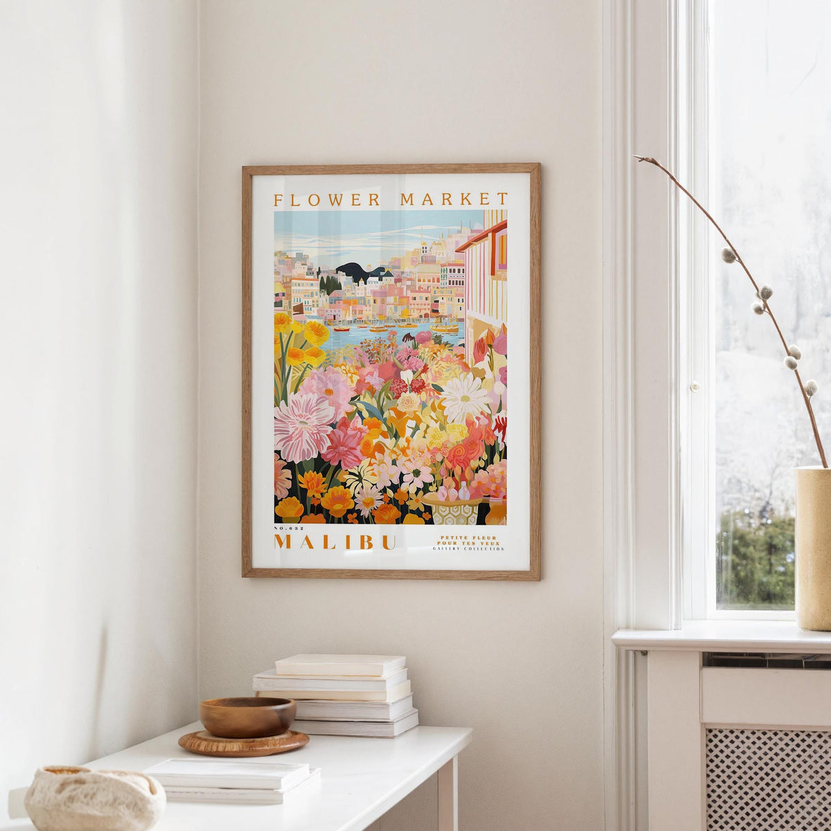 a framed poster of a colorful floral scene hanging on a wall, with a white table and a vase of flowers in the foreground.