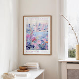 a framed poster of a cityscape with a floral design, hanging on a wall in a room with a white table and a window.