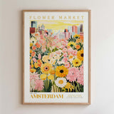 The image is a framed poster depicting a vibrant and colorful flower market in Amsterdam, with a variety of flowers and plants in the foreground and a cityscape in the background.
