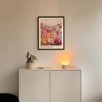 a white cabinet with a potted plant and a lamp on top, set against a wall with a framed poster of a flower market above it.