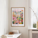 a framed poster of a colorful cityscape with flowers hanging on a wall in a room with a white table and a window.