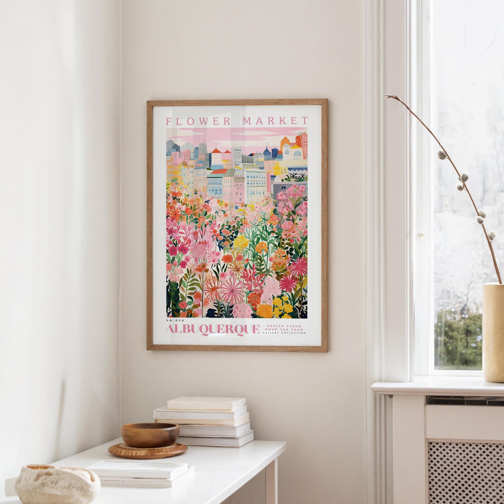 a framed poster of a colorful cityscape with flowers hanging on a wall in a room with a white table and a window.