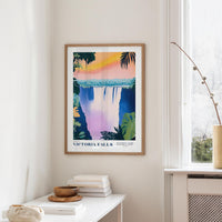 a framed poster of Victoria Falls hanging on a wall, with a white table and a plant in the foreground.