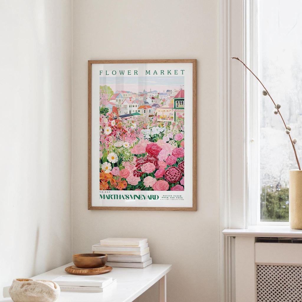 a framed poster of a colorful garden scene hanging on a wall, with a white table and a vase of flowers in the foreground.