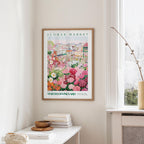 a framed poster of a colorful garden scene hanging on a wall, with a white table and a vase of flowers in the foreground.