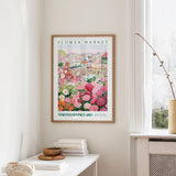a framed poster of a colorful garden scene hanging on a wall, with a white table and a vase of flowers in the foreground.