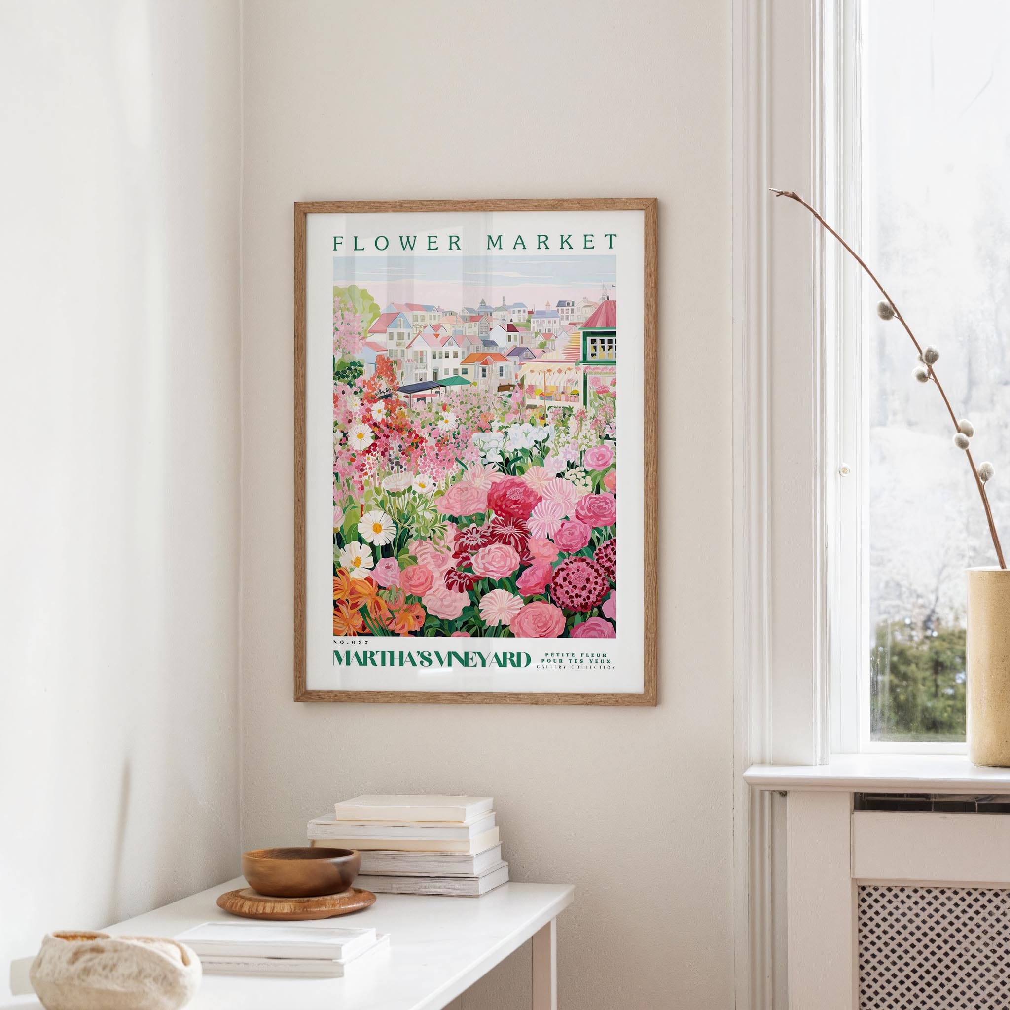 a framed poster of a colorful garden scene hanging on a wall, with a white table and a vase of flowers in the foreground.