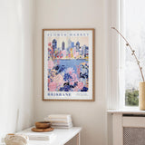 a framed poster of a cityscape with a floral design, hanging on a wall in a room with a window and a small table with books and a bowl on it.