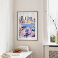 a framed poster of a cityscape with a floral design, hanging on a wall in a room with a window and a small table with books and a bowl on it.