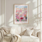 a framed poster of a colorful floral scene hanging on a wall above a white couch with pillows.
