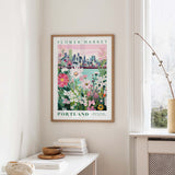 a framed poster of a cityscape with a flower market in Portland, Oregon, hanging on a wall.