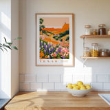 a framed poster of a colorful landscape of flowers hanging on a wall above a wooden countertop with a bowl of lemons.