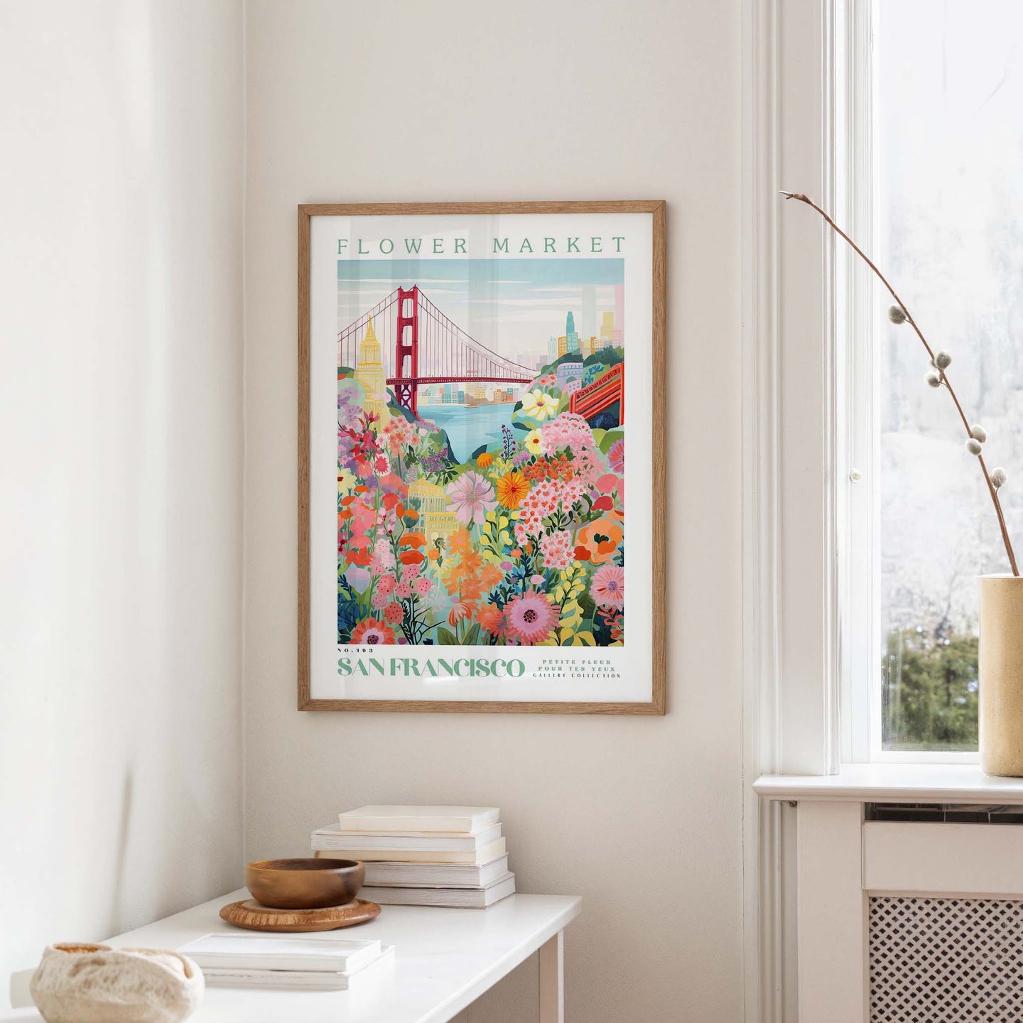 a framed poster of a colorful, floral-themed illustration of the Golden Gate Bridge in San Francisco, hanging on a wall in a room with a window and a small table with books and a vase.