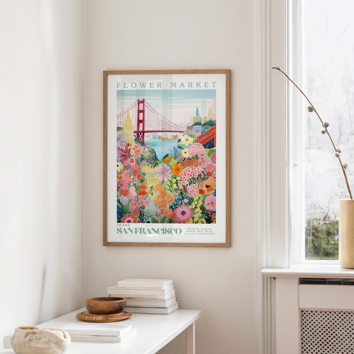 a framed poster of a colorful, floral-themed illustration of the Golden Gate Bridge in San Francisco, hanging on a wall in a room with a window and a small table with books and a vase.
