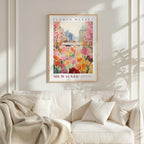 a framed poster of a colorful floral scene hanging on a wall above a white couch with a blanket draped over it.