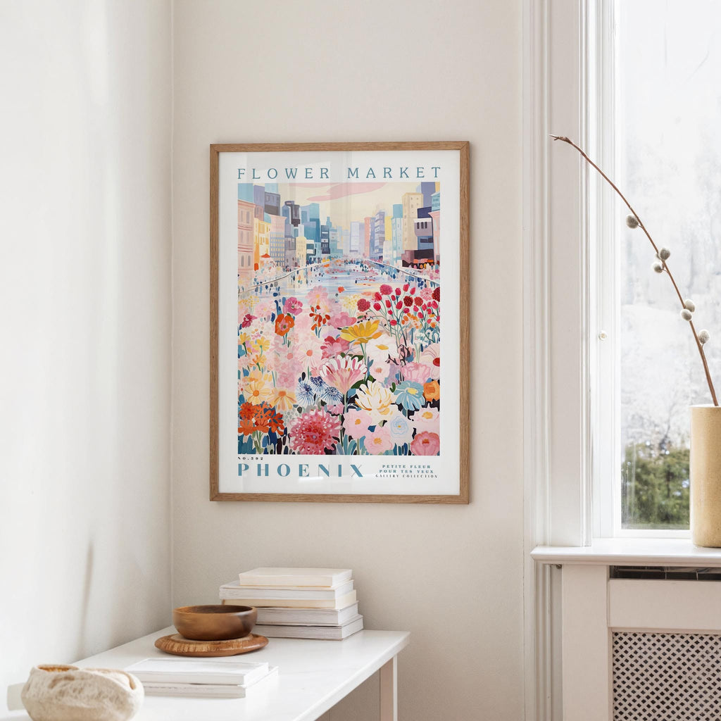 a framed poster of a colorful cityscape with flowers hanging on a wall, next to a window and a small table with books and a bowl.