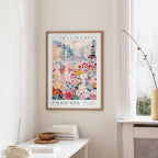 a framed poster of a colorful cityscape with flowers hanging on a wall, next to a window and a small table with books and a bowl.