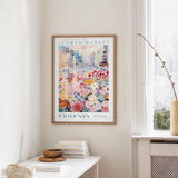 a framed poster of a colorful cityscape with flowers hanging on a wall, next to a window and a small table with books and a bowl.