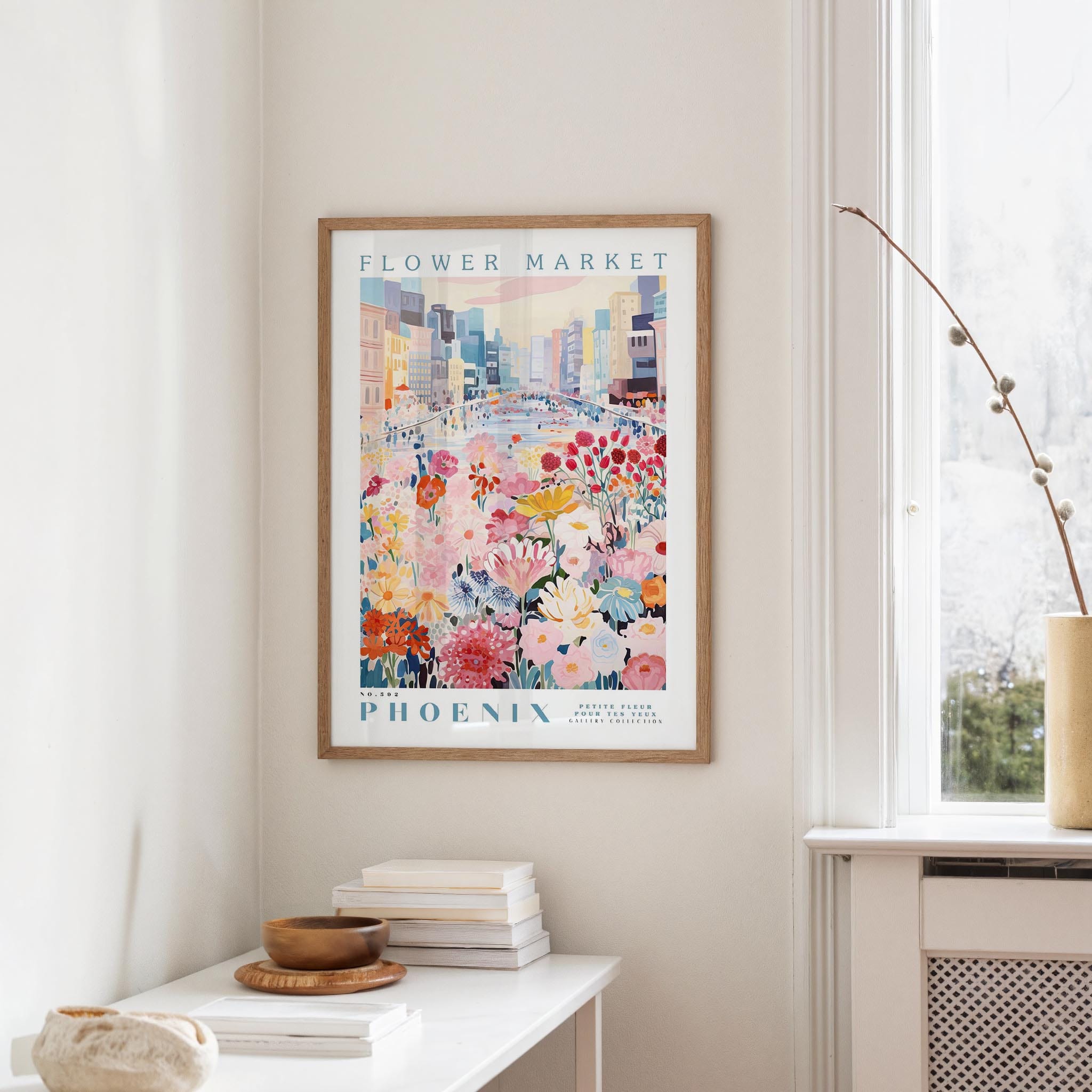 a framed poster of a colorful cityscape with flowers hanging on a wall, next to a window and a small table with books and a bowl.