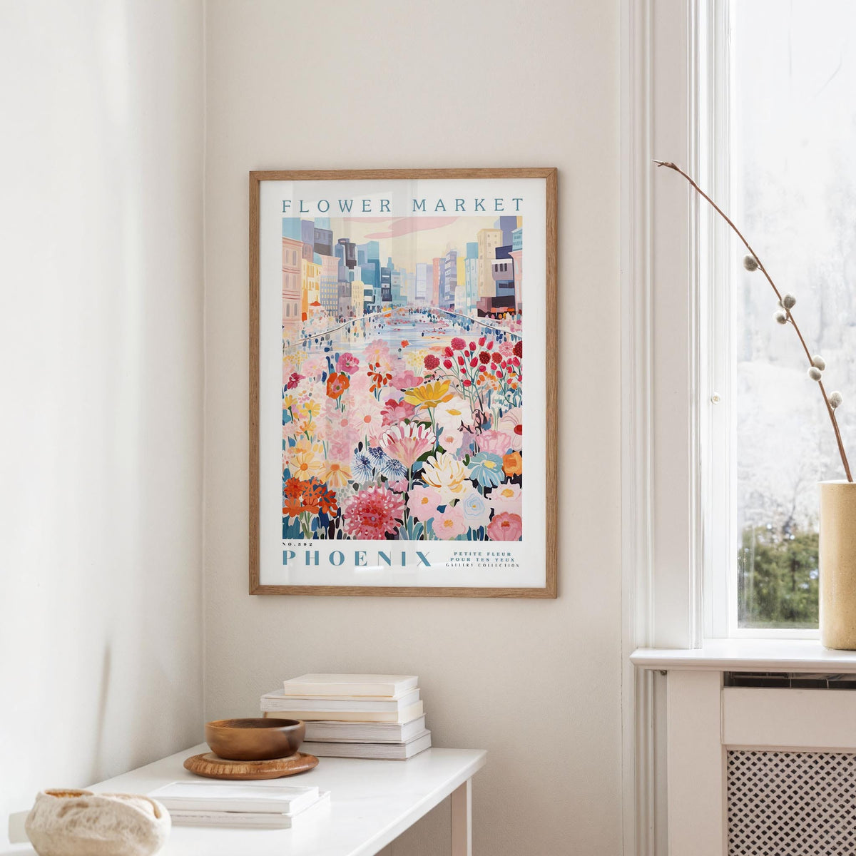 a framed poster of a colorful cityscape with flowers hanging on a wall, next to a window and a small table with books and a bowl.