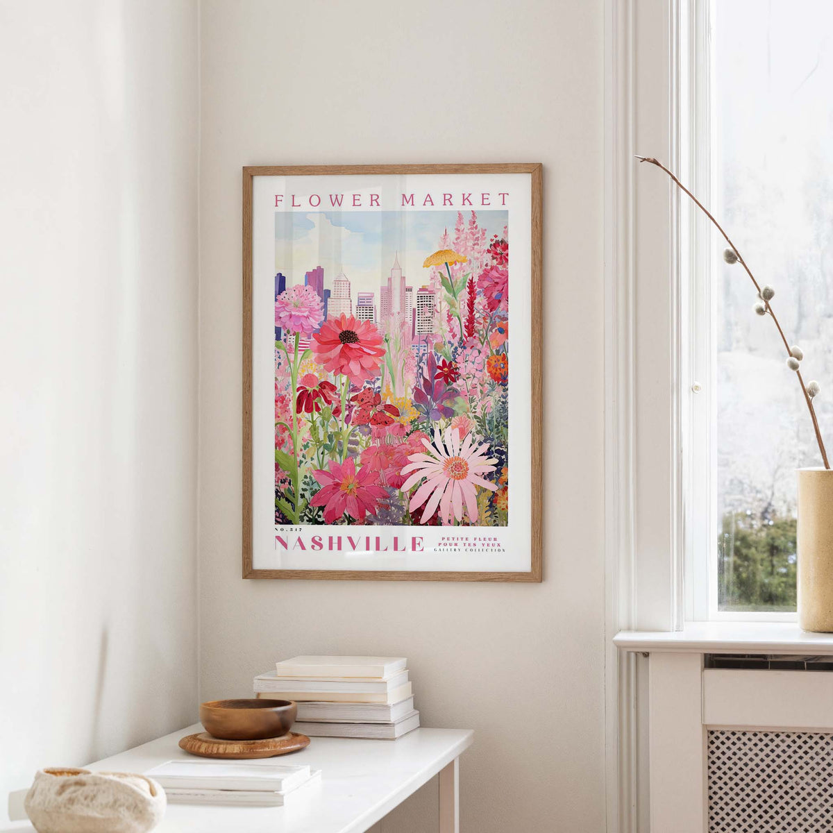 a framed poster of a colorful floral scene hanging on a wall, with a window and a small table with books and a vase in the foreground.
