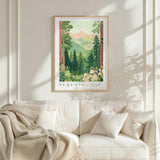 a framed poster of a serene forest scene with mountains in the background, hanging on a wall above a white couch with a blanket draped over it.