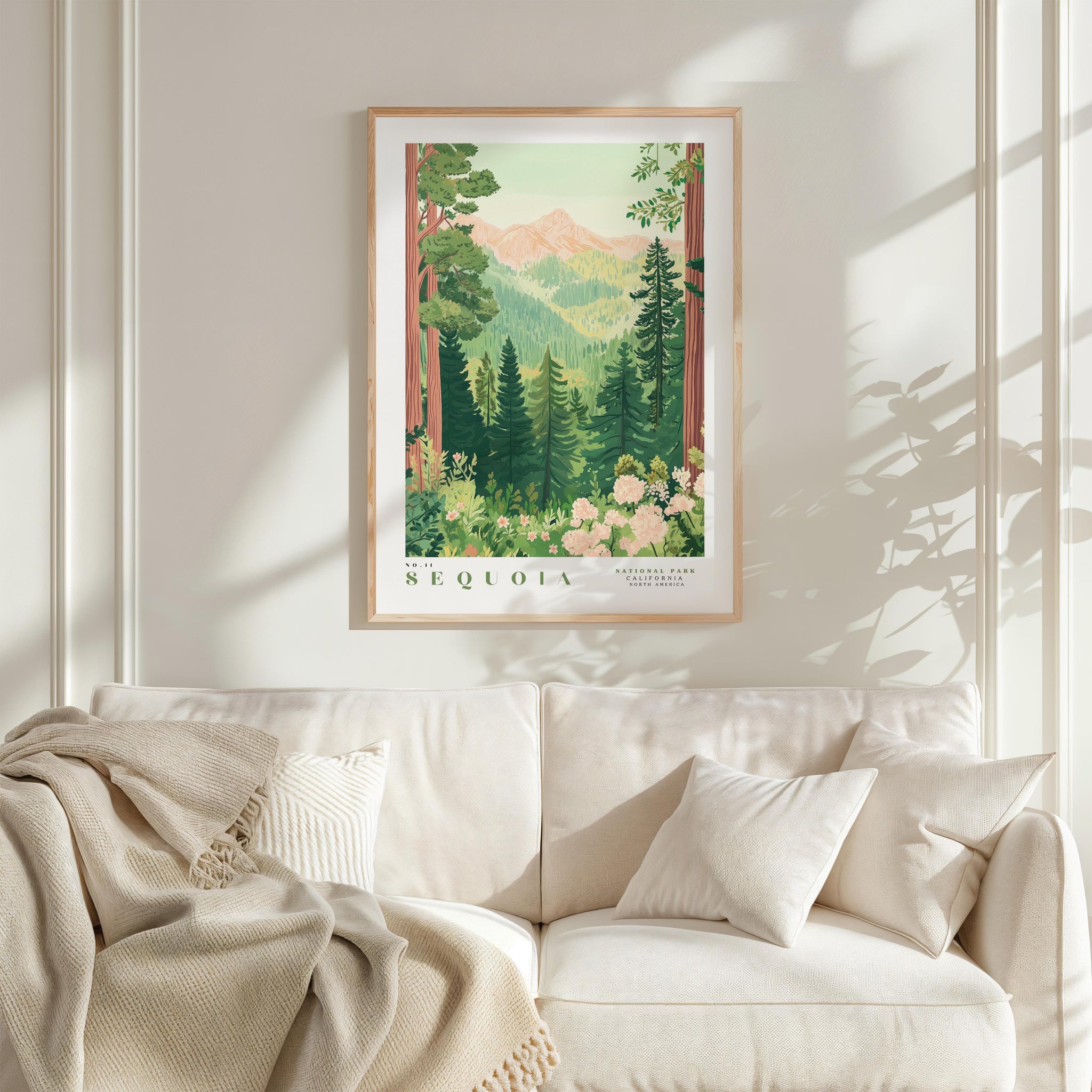 a framed poster of a serene forest scene with mountains in the background, hanging on a wall above a white couch with a blanket draped over it.