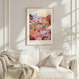 a framed poster of a colorful cityscape with a flower market in the background, hanging on a wall above a white couch with a blanket draped over it.