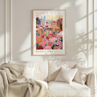a framed poster of a colorful cityscape with a flower market in the background, hanging on a wall above a white couch with a blanket draped over it.