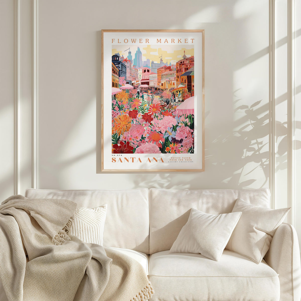 a framed poster of a colorful cityscape with a flower market in the background, hanging on a wall above a white couch with a blanket draped over it.