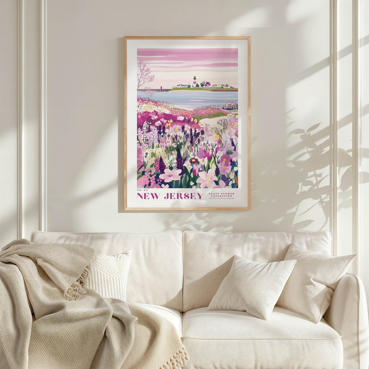 a framed poster of a scenic landscape with a field of flowers and a body of water, hanging on a wall above a white couch with a blanket draped over it.