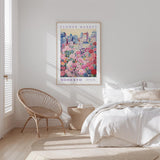 a bedroom with a large poster of a flower market hanging on the wall, a woven chair, and a bed with white bedding.
