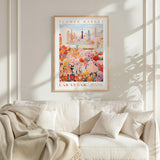 a framed poster of a cityscape with a flower market in the background, hanging on a wall above a white couch with pillows.