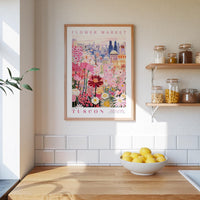 a framed poster of a colorful floral scene hanging on a wall above a kitchen counter with a bowl of lemons on it.