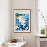 a framed poster of a beach scene with palm trees, flowers, and a blue sky, hanging on a wall in a room with a white table and a window.