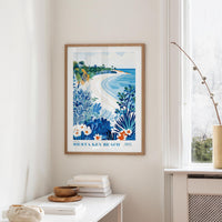a framed poster of a beach scene with palm trees, flowers, and a blue sky, hanging on a wall in a room with a white table and a window.