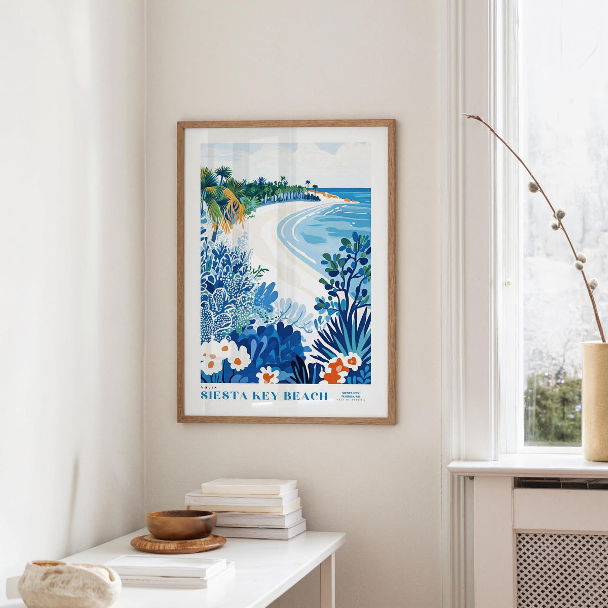 a framed poster of a beach scene with palm trees, flowers, and a blue sky, hanging on a wall in a room with a white table and a window.