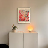 a white cabinet with a potted plant and a lamp on top, and a framed poster of a flower market hanging on the wall above it.