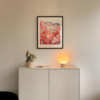 a white cabinet with a potted plant and a lamp on top, and a framed poster of a flower market hanging on the wall above it.
