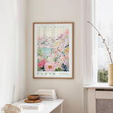 a framed poster of a colorful floral scene hanging on a wall, with a window and a small table with books and a vase nearby.