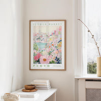 a framed poster of a colorful floral scene hanging on a wall, with a window and a small table with books and a vase nearby.