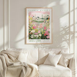 A framed poster of a flower market in Adelaide, Australia, hangs on a white wall above a beige couch with a blanket draped over it.