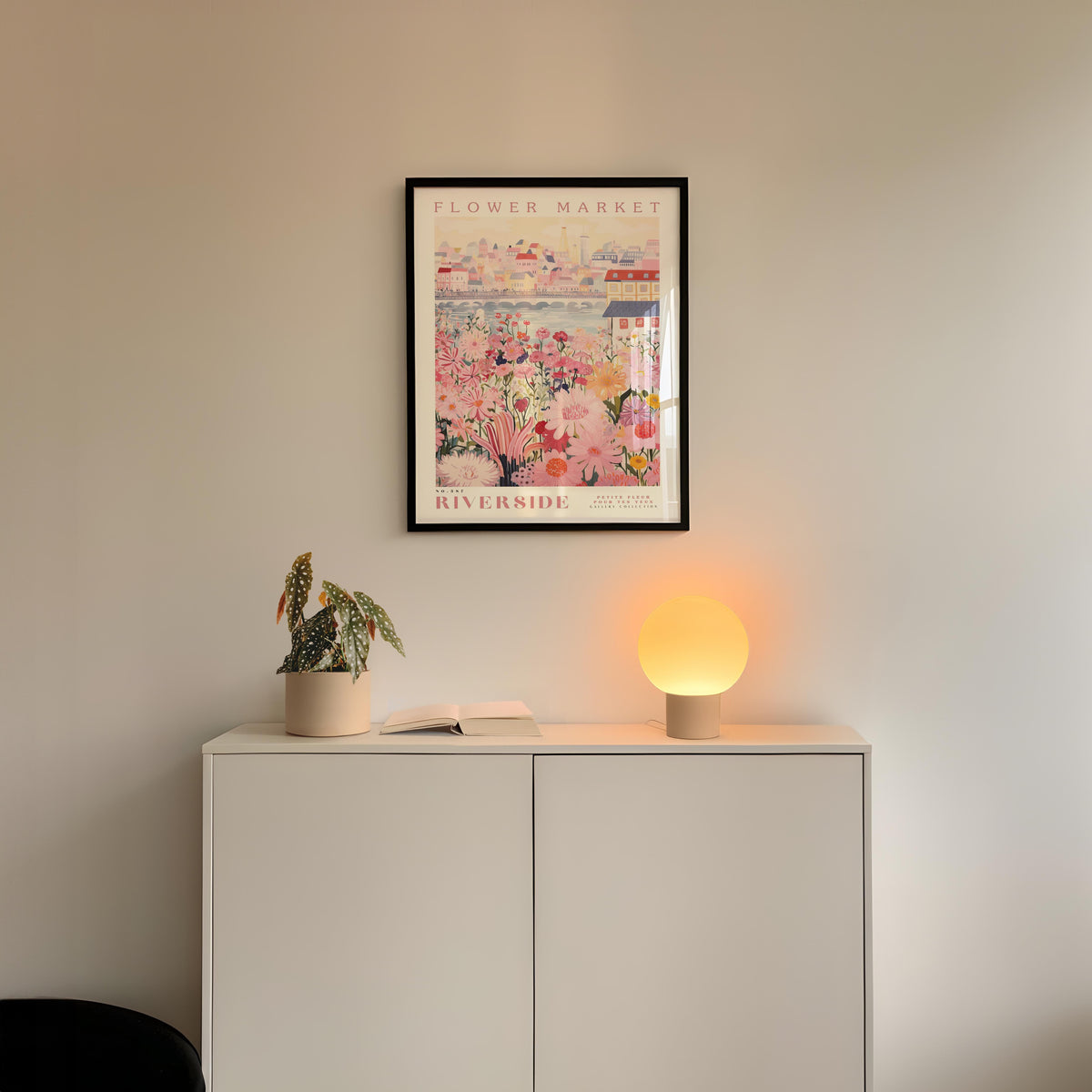 a white cabinet with a potted plant and a lamp on top, set against a white wall with a framed poster of a flower market above it.